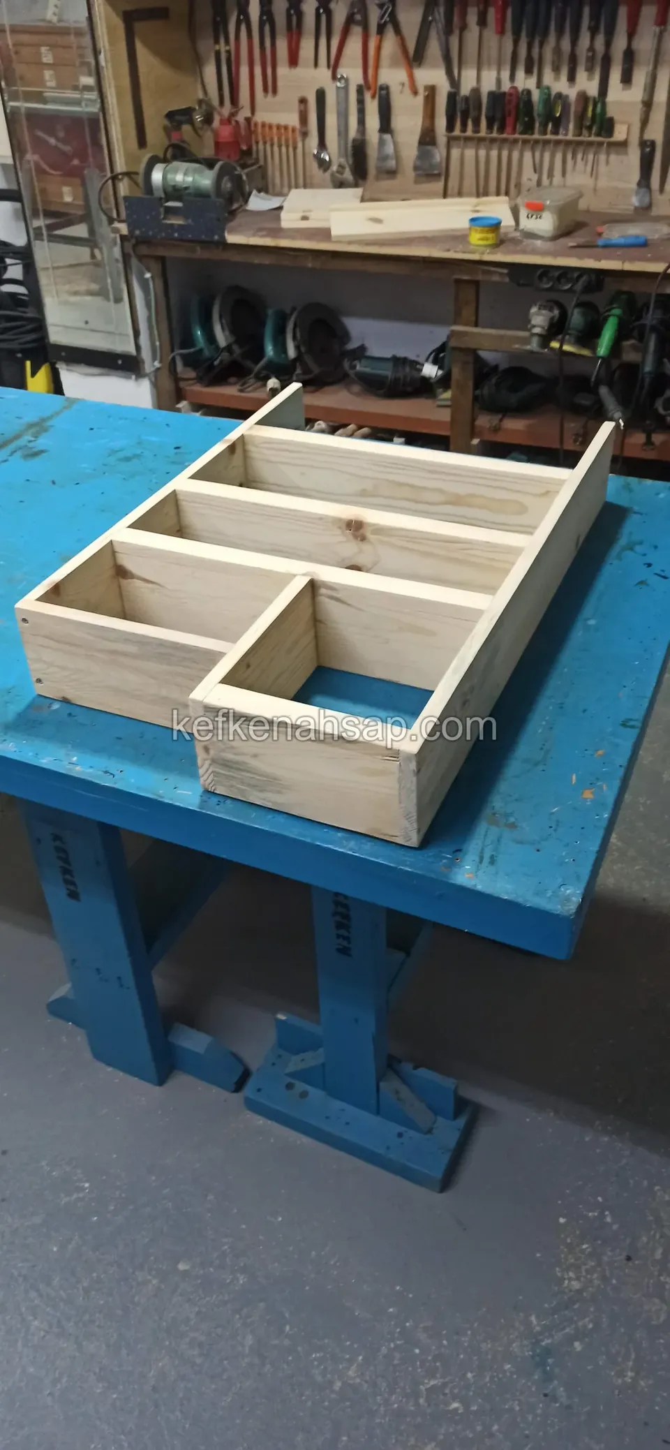Ein helles Holzregalgerüst mit Fächern flach auf einer blauen Werkbank, an der Wand dahinter Handwerkzeug