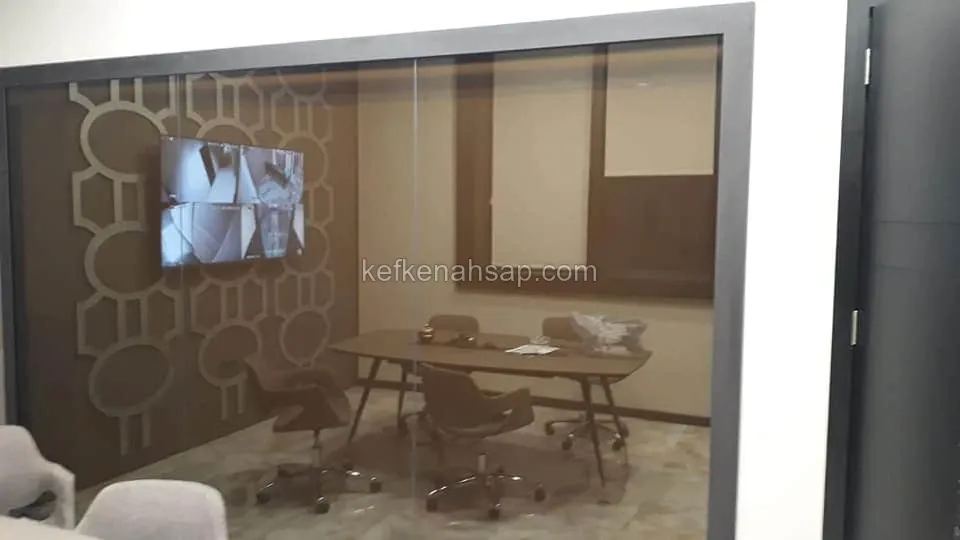 Cam bölmeli toplantı odasında oval masa, tekerlekli koltuklar ve ekranın bulunduğu geometrik desenli kahverengi duvar paneli