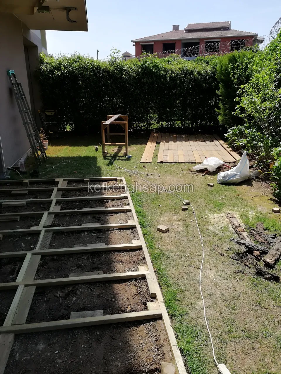 Timber deck joist frame on soil with lawn, tools and the first laid boards beside it