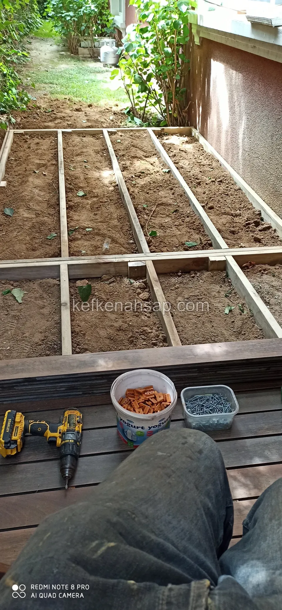 Deck sub-joists laid over bare soil along a wall, with a drill and screw tubs in front