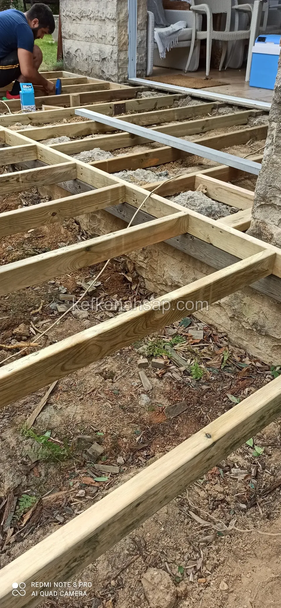 Network of deck joists laid on soil, with a worker at the top corner and a metal profile reaching the threshold