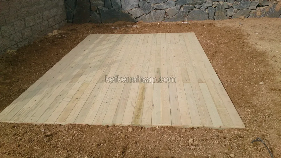 Toprak zeminde yatan, tahtaları boyuna döşenmiş yeni açık renkli dikdörtgen deck; arkada taş duvar