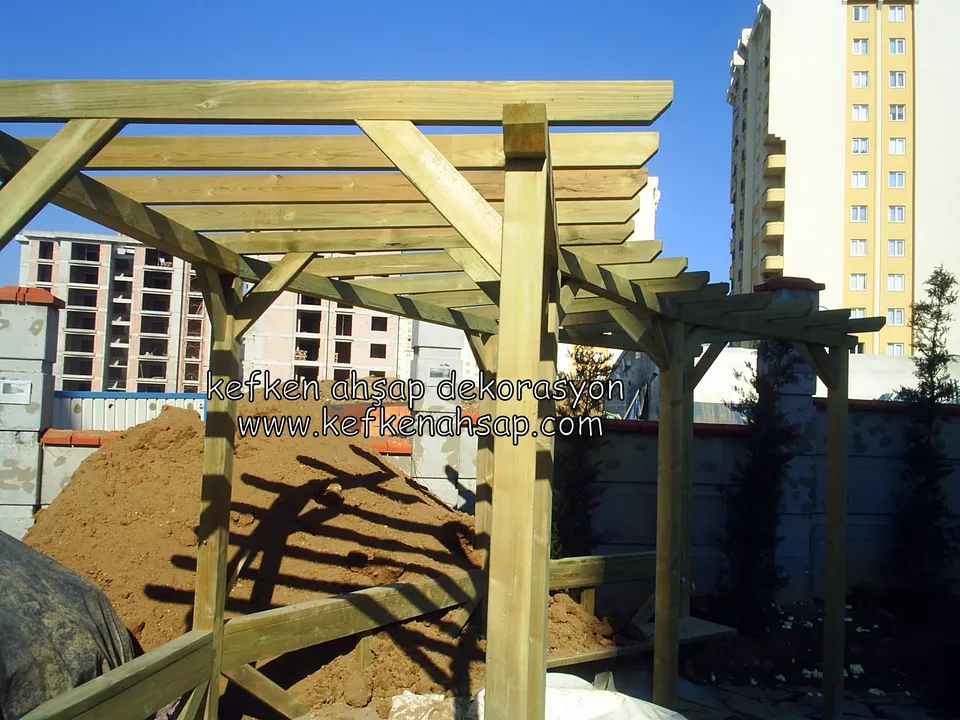 Toprak yığınının yanında yükselen ahşap pergola iskeleti, payandalı kirişler ve arkada çok katlı apartmanlar