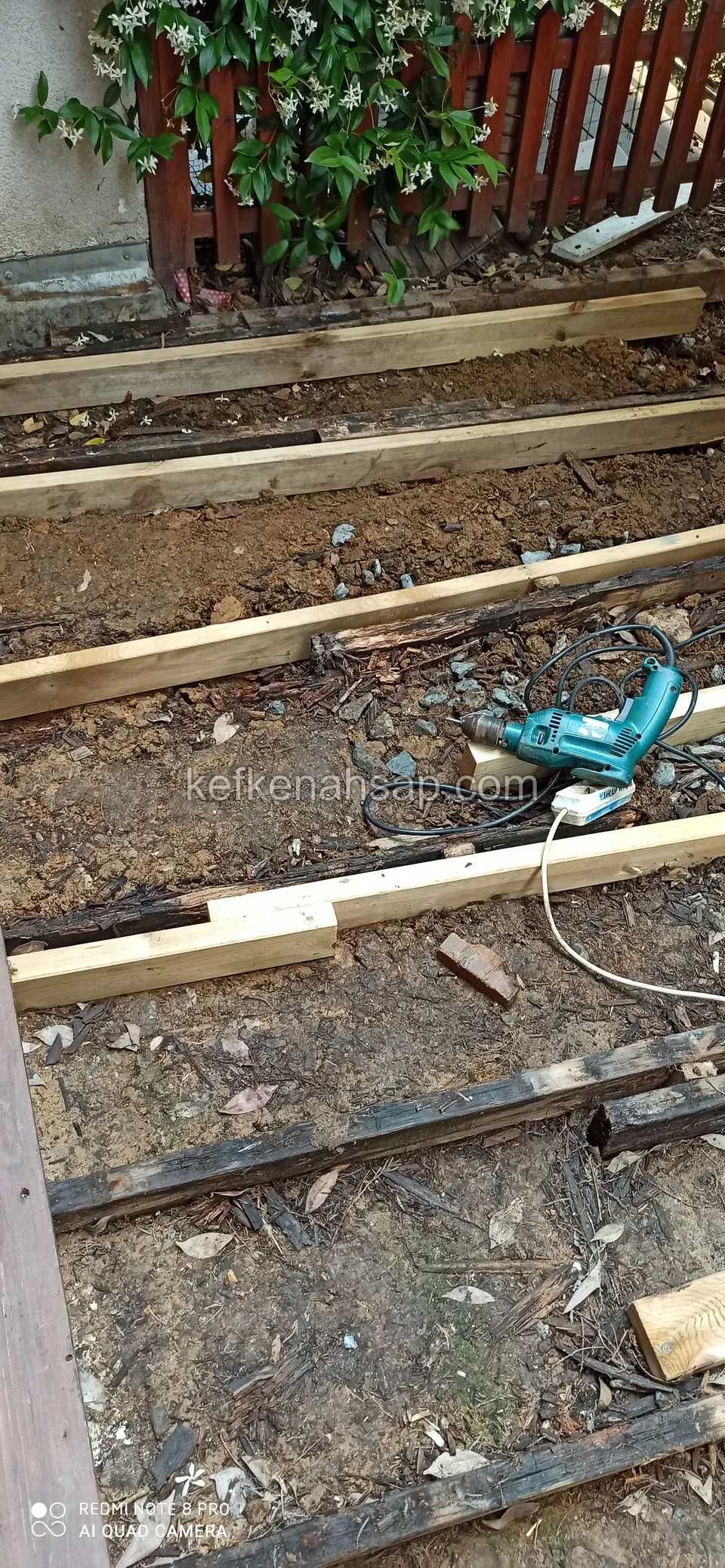 Timber joists set on bare ground with a drill lying beside them, and a garden fence behind