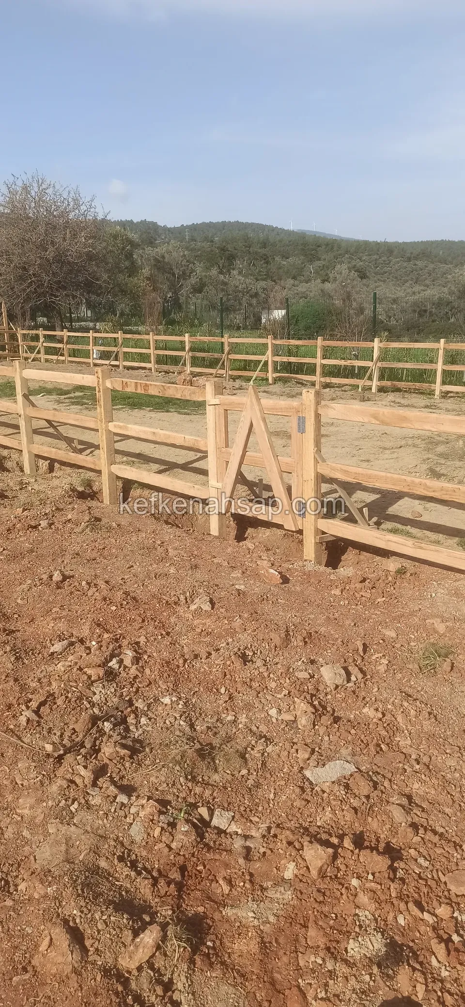 Kırmızı toprak zeminde ahşap yatay çıtalı çit hattı ve içinde çaprazlı kapı çerçevesi, uzakta ağaçlı tepe