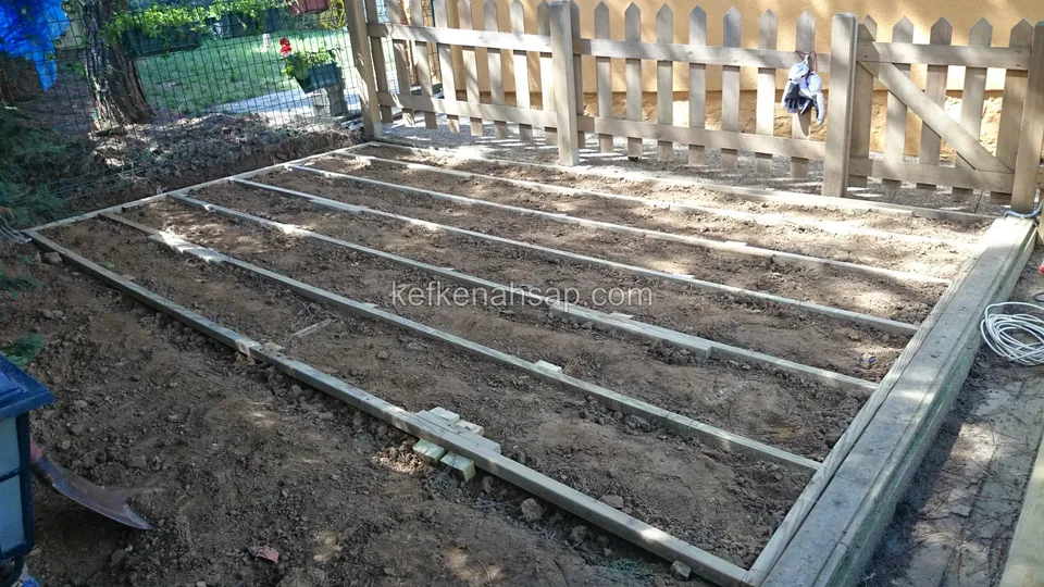 Toprak üzerine kurulmuş ahşap çerçeve ve içindeki ince kirişler; arkada kapılı ahşap çit, sağda elektrik kablosu