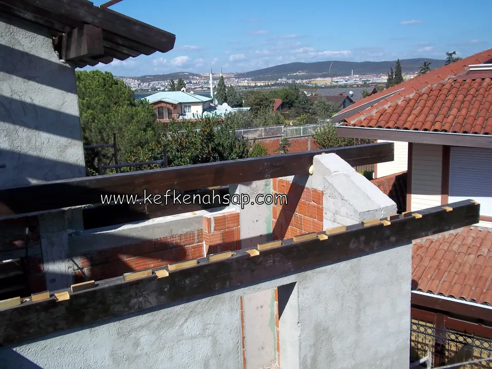 Tuğla ve beton duvarın üstüne dizilmiş koyu renk ahşap kirişler, arkada kiremit çatılı evler ve tepeler