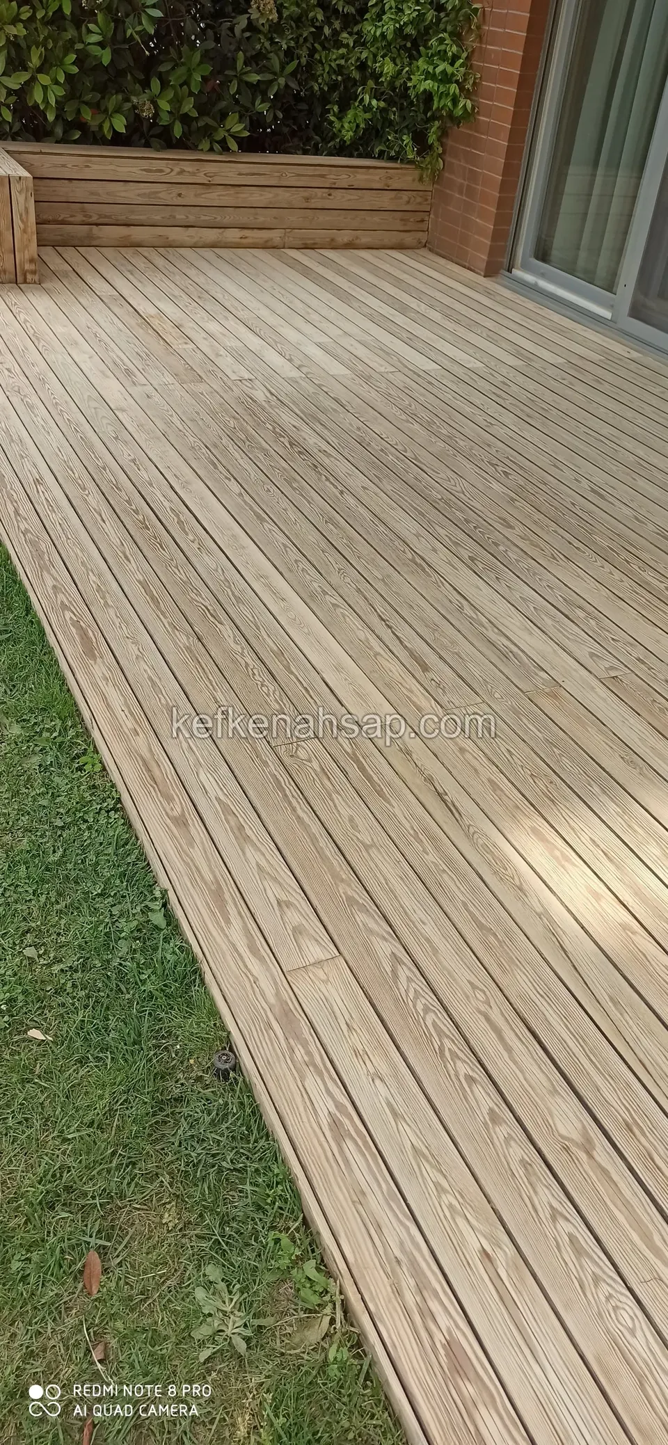 Unbehandeltes helles Kiefern-Deck am Rasenrand vor einer Backsteinwand, dahinter eine Holzbank