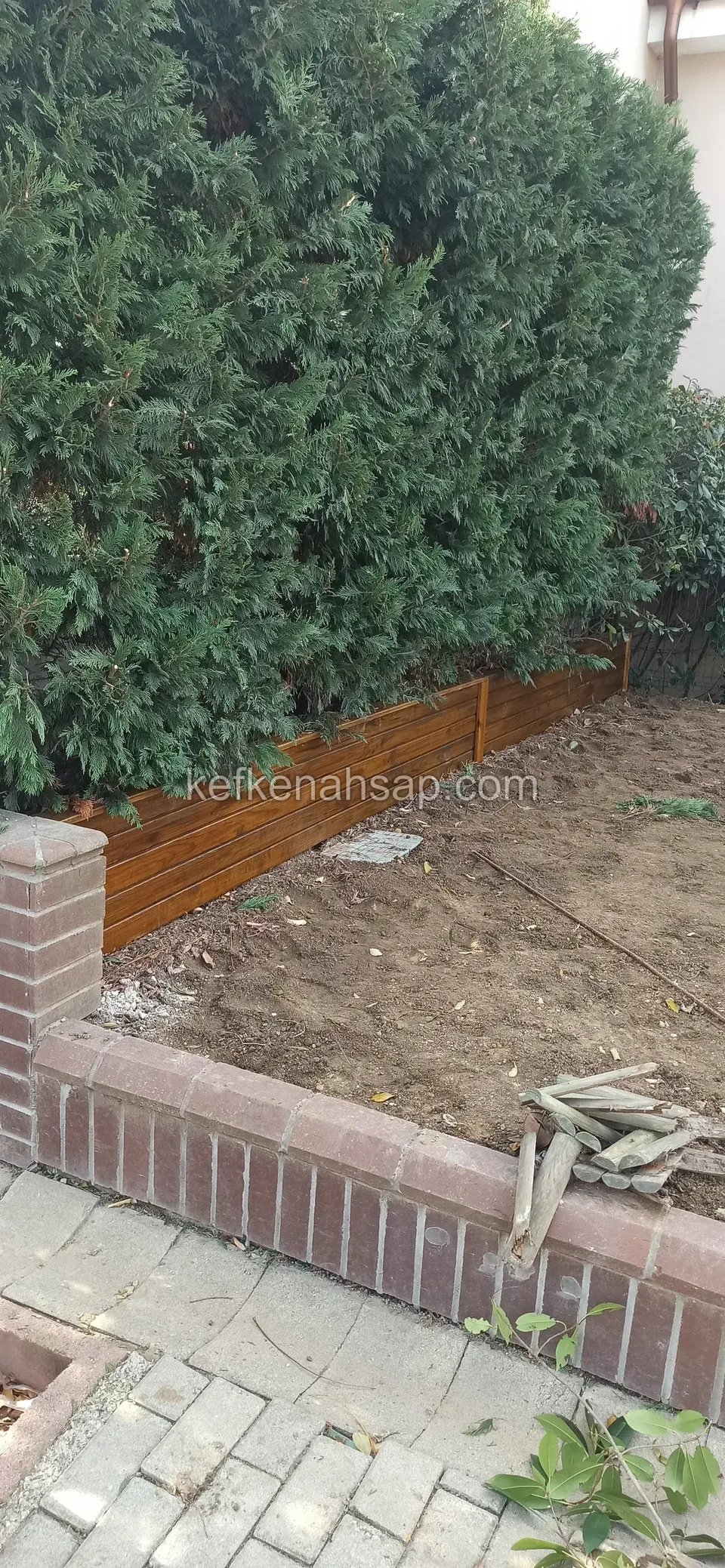 Tuğla kenarlı toprak bahçe yatağının arkasında ahşap kenarlık, yerde kalıntı parçalar, üstte yeşil çalılık