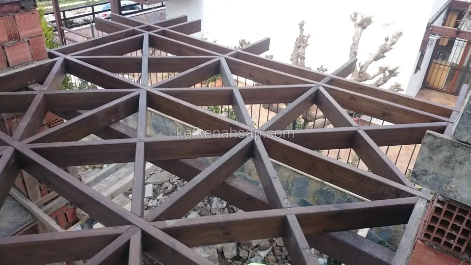 Tuğla kolon ve taş duvar arasında geometrik üçgen desenli vernikli pergola çatı kafesi, arkada korkuluk ve ev