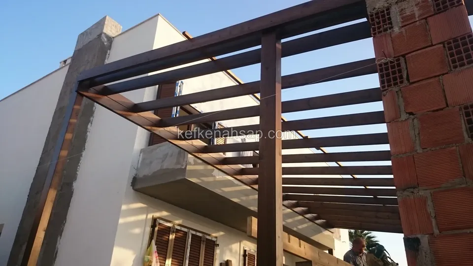 Binanın yanında tuğla kolona oturan vernikli pergola çerçevesi ve sıralı kirişler, alt katta panjurlu pencereler