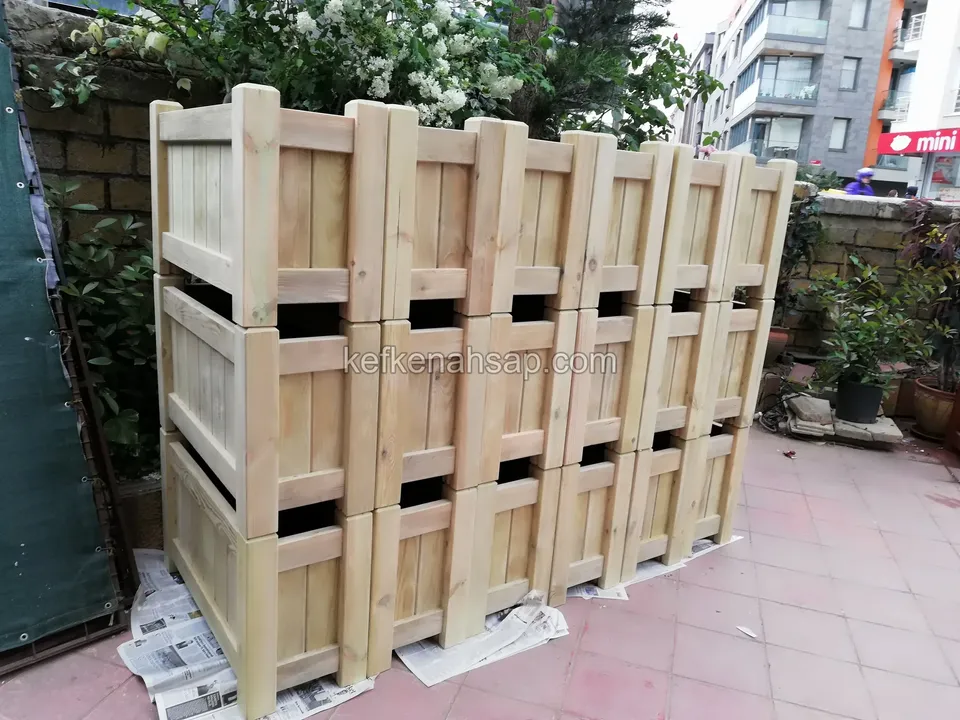 Teras zemininde üç sıra hâlinde üst üste yığılmış çok sayıda açık renk ahşap çiçeklik kutusu