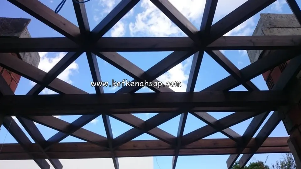 Alttan görünen koyu renkli ahşap pergola kirişleri, üçgen desen oluşturan çaprazlar ve aralarından görünen mavi gökyüzü