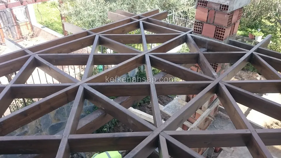 Üçgen geometrik desenli pergola kirişleri, altta bahçe, zeytin ağaçları ve inşaat artıkları, köşede tuğla kolon