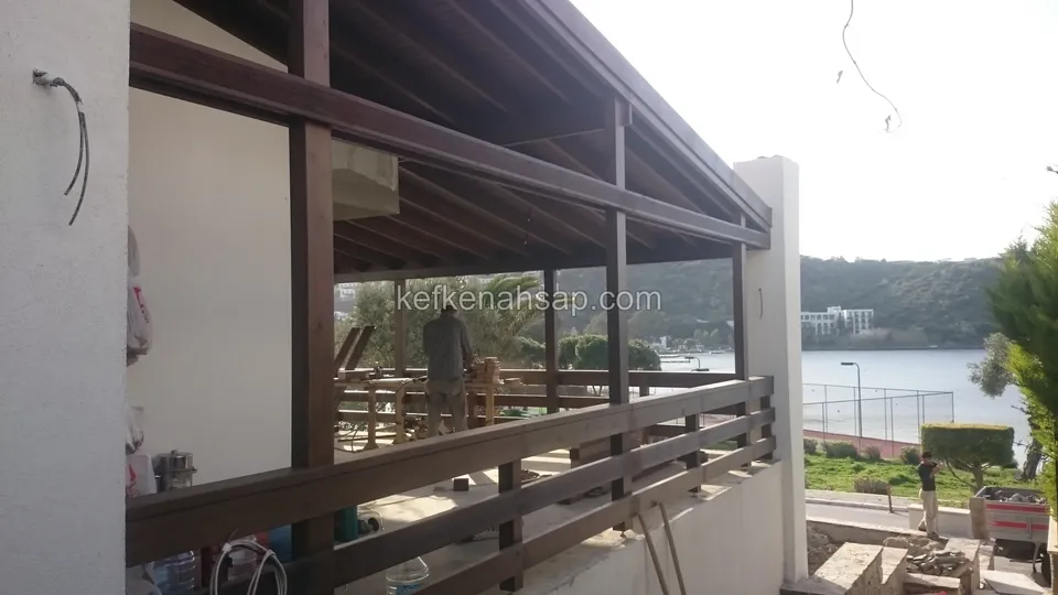 Üst kat terasında koyu ahşap korkuluk ve çatı; arkada çalışan işçiler ve koy görünüyor