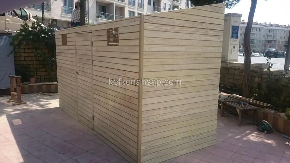 Apartmanlar önünde, köşeden görünen yatay tahtalı uzun ahşap kasa; üst kenarda iki küçük kare menfez var