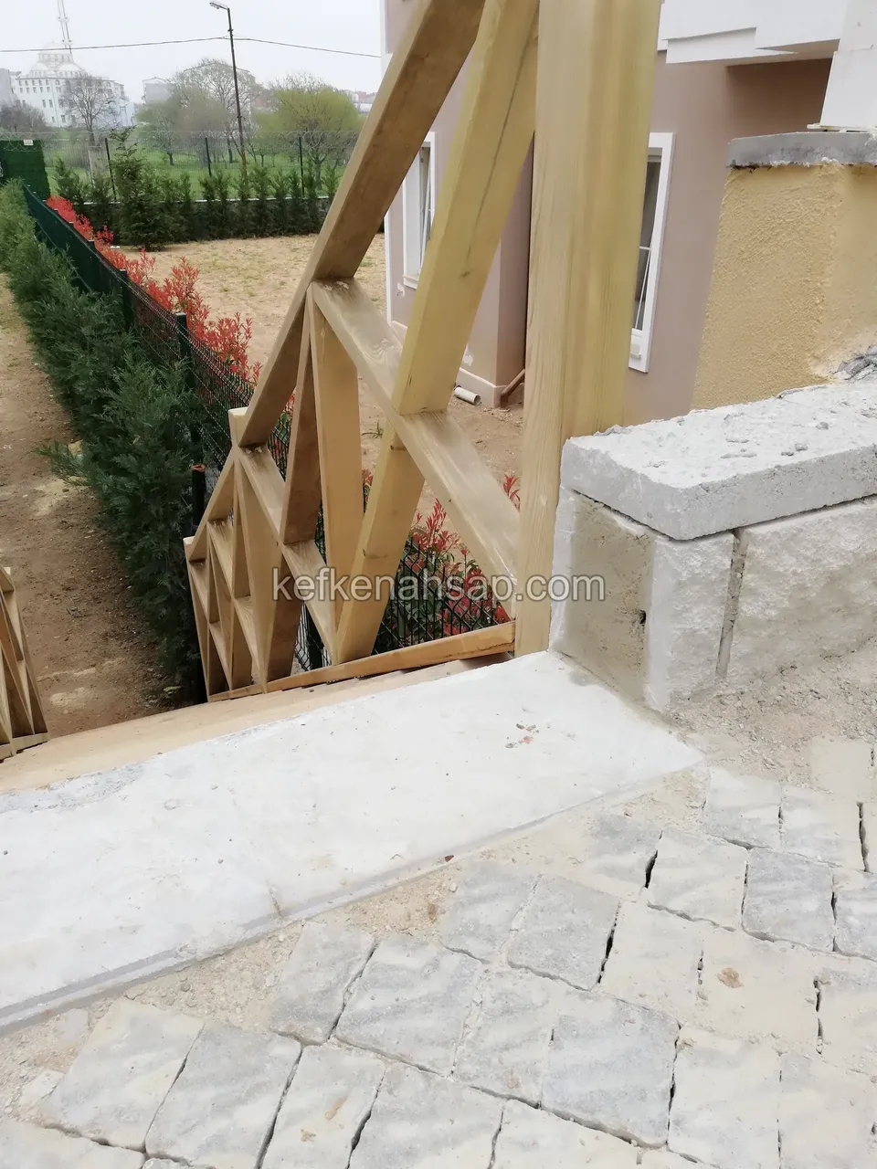 Blick von oben auf Holztreppe, Kreuzstreben-Geländer und Steinpflaster, seitlich Zypressenhecken