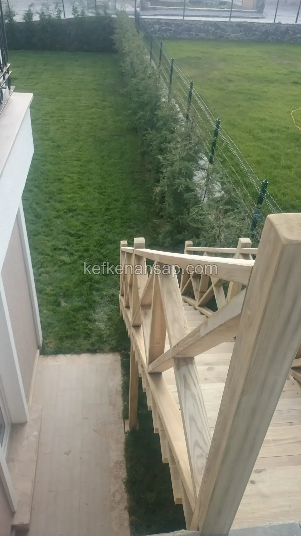 Üstten görünen ahşap merdiven korkuluğu, kalın başlık direği, aşağıda çimlik, servi sırası ve tel çit