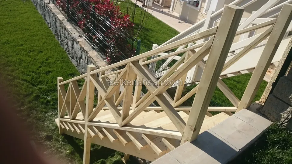 Üstten görünen ahşap merdiven ve çaprazlı korkuluk; yanda taş duvar, kırmızı çalılar ve çimlik
