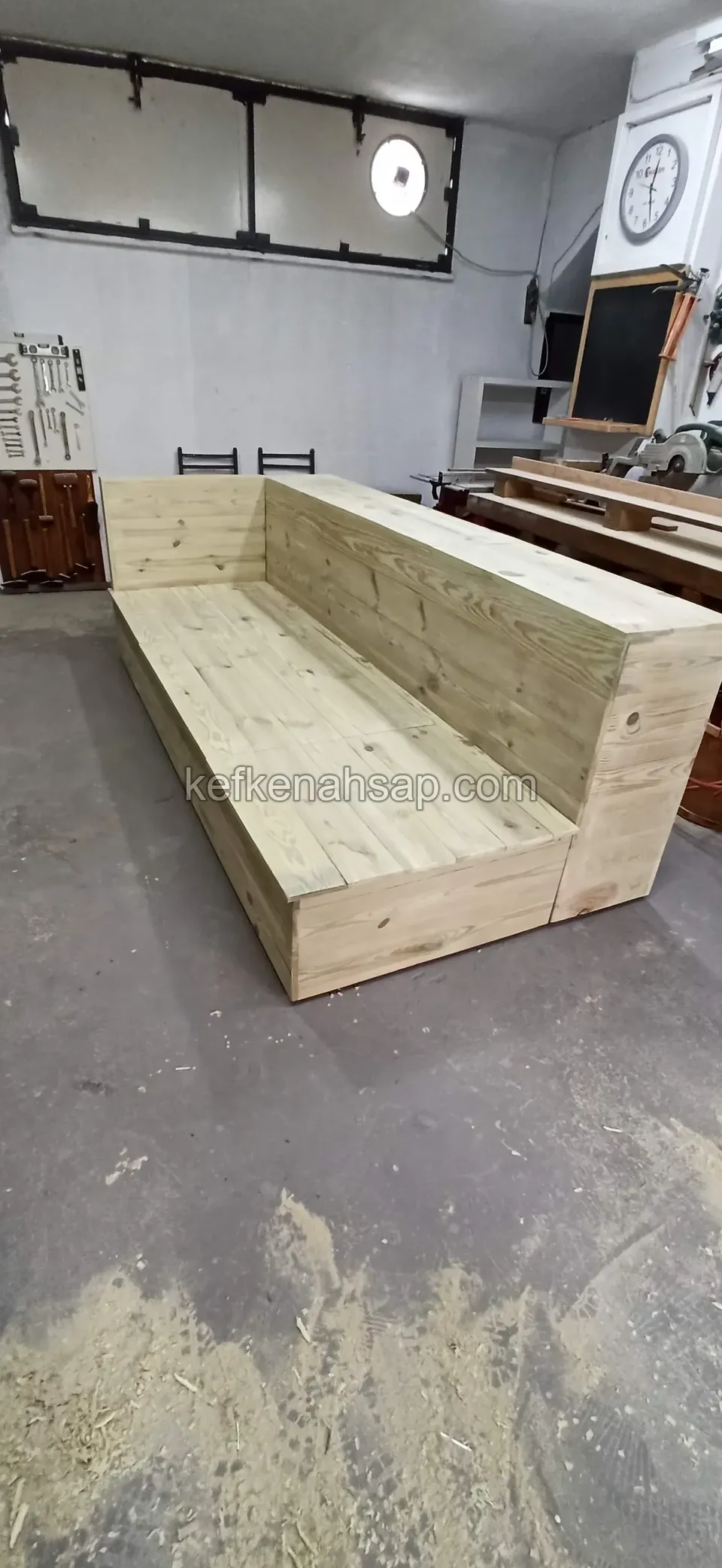 Eine lange Holzsitzeinheit mit hoher Lehne aus hellgrünen Kiefernbrettern auf dem grauen Boden einer Werkstatt