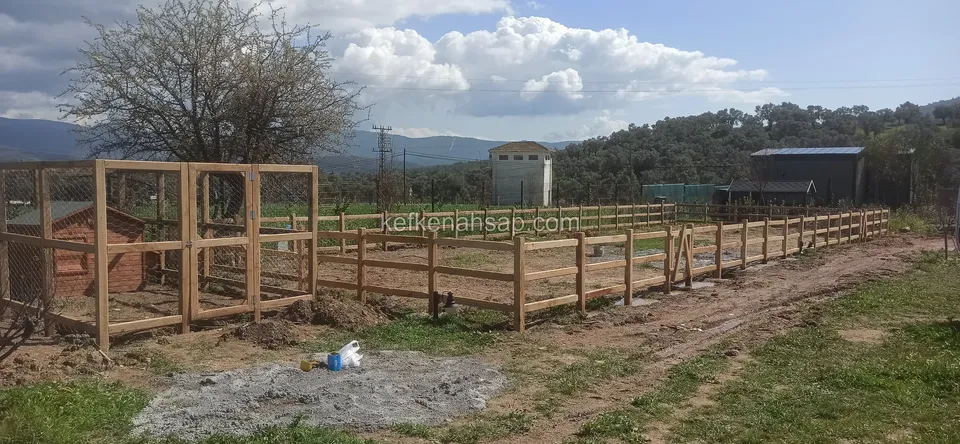 Açık arazide uzanan yatay çıtalı çit hattı ve başında iki kapı çerçevesi, yanında kulübe ve uzakta dağlar