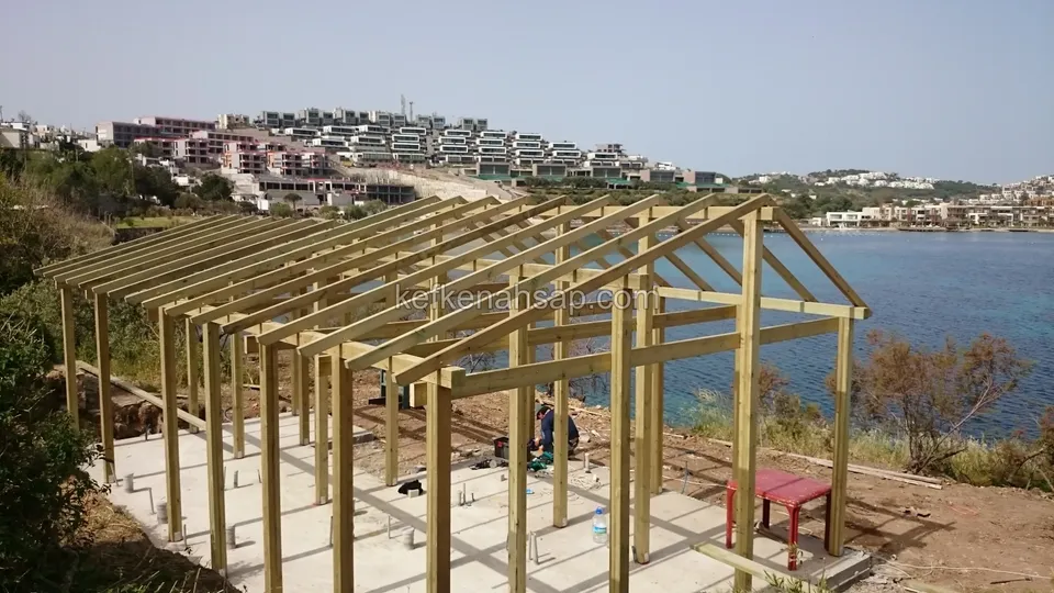 Uzaktan deniz kenarındaki ahşap kamelya iskeleti, beton zeminde kırmızı bir sandalye, arkada yamaç evleri