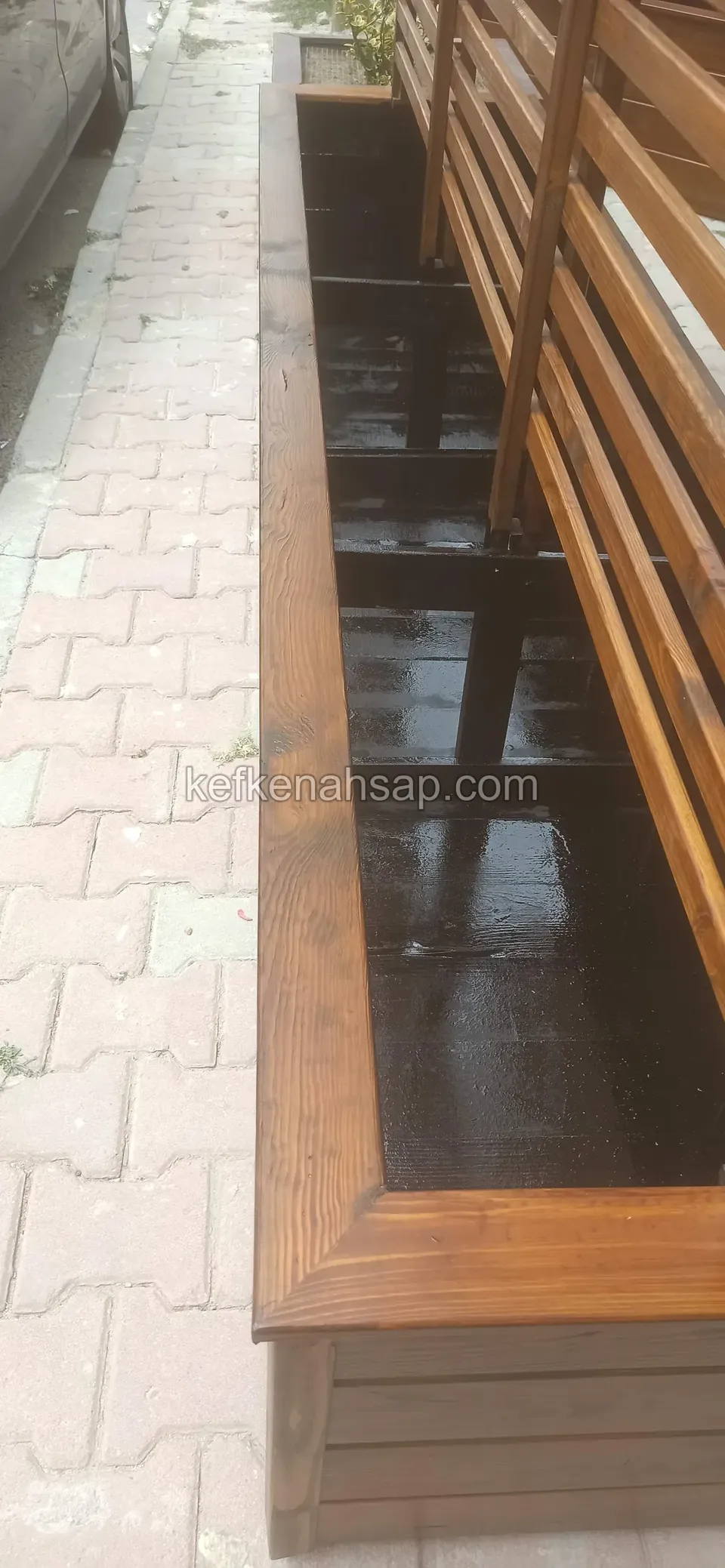 Close view of the edge profile of a long timber planter on the pavement and the lower part of the slatted screen above it