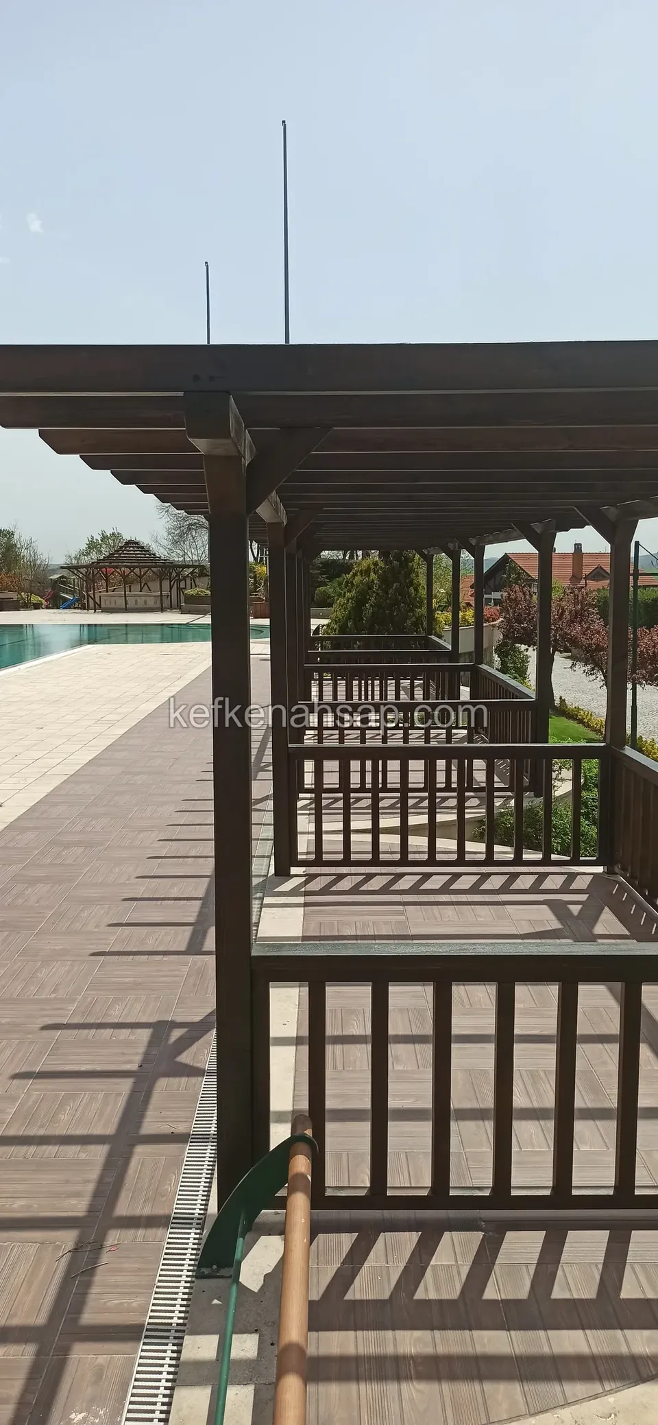 Uzun yol boyunca sıralanan korkuluklu vernikli pergola, uzakta havuz ve yanında lamba direkleri