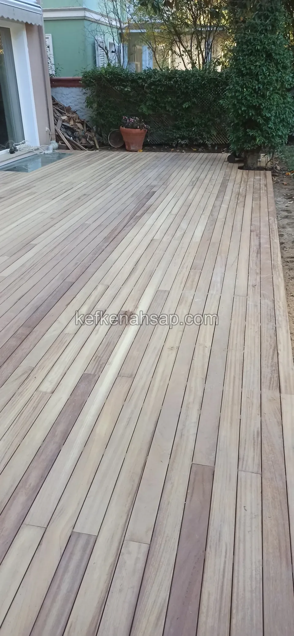Light yellowish-brown deck boards laid long and side by side, with a glass door and hedge alongside