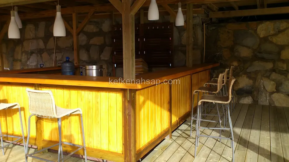 Taş duvar ve kirişli çatı altında vernikli ahşap bar tezgahı; önünde yüksek tabureler, üstte sarkıt lambalar