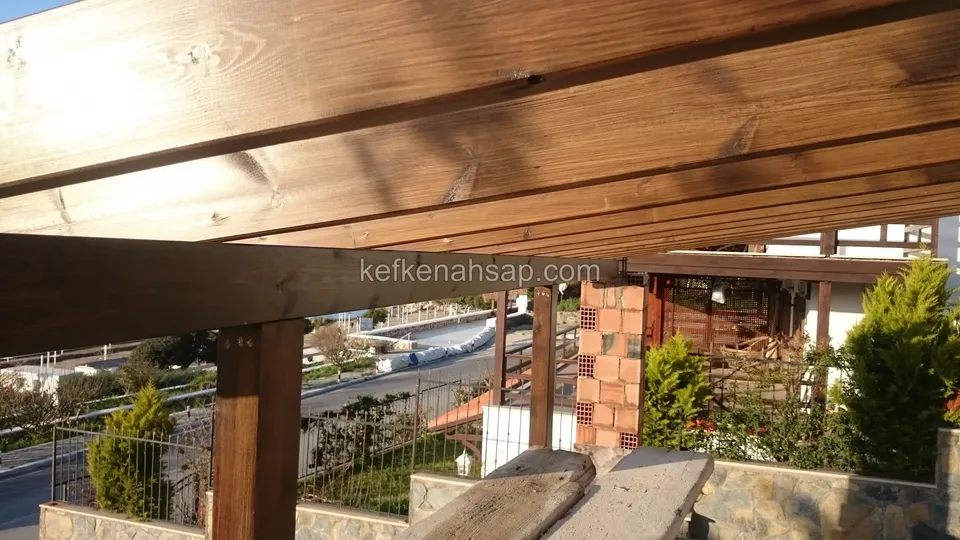 Yakından vernikli kalın pergola kirişi ve direği, arkada tuğla kolon, bahçe ve kıvrılan yol