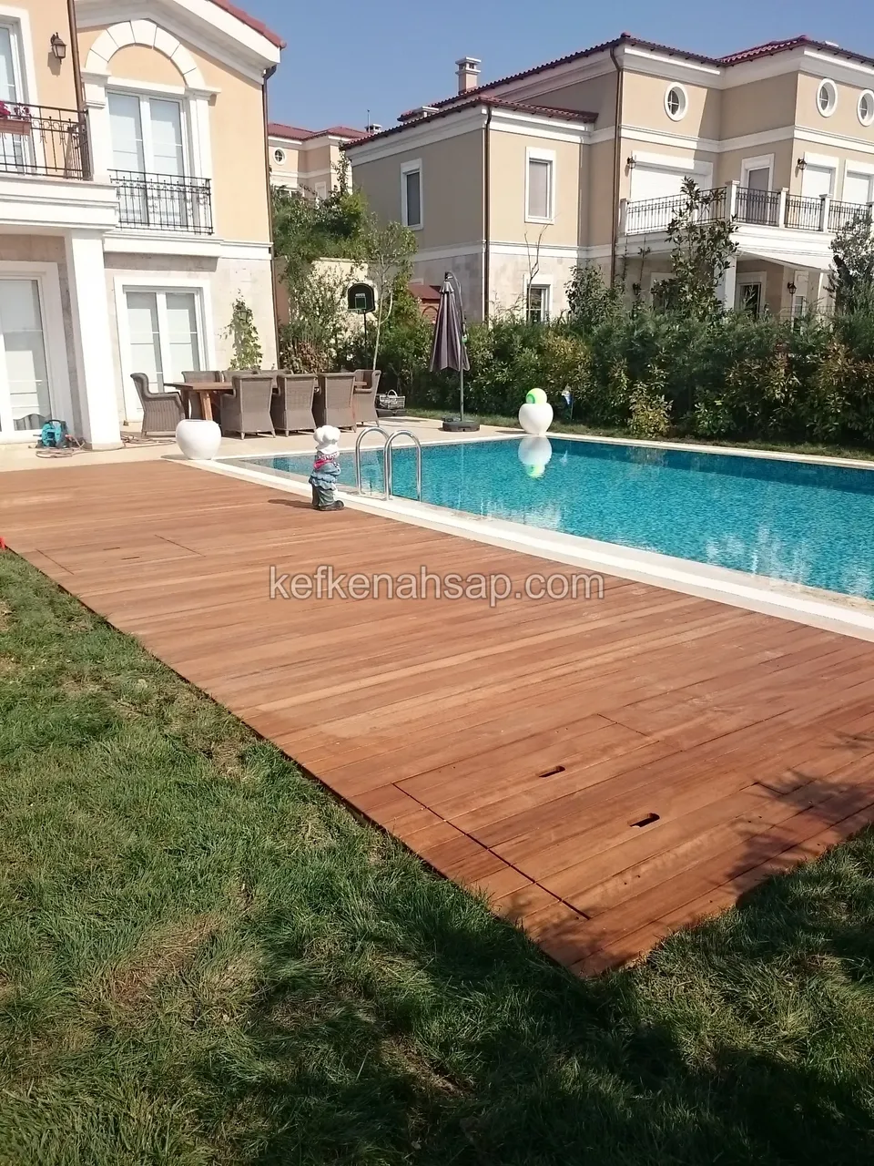 Havuz kenarında çimlere doğru açılan turuncu kahve deck, arkada sarı boyalı iki katlı evler ve masa takımı