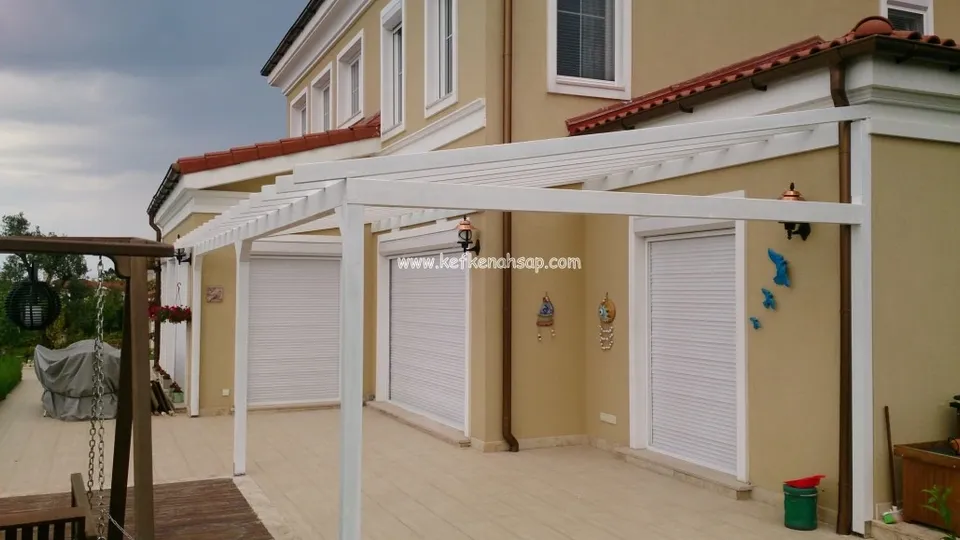 Kiremit çatılı, sarı cepheli bir villanın teras boyunca uzanan, duvara bitişik beyaz boyalı ahşap pergola