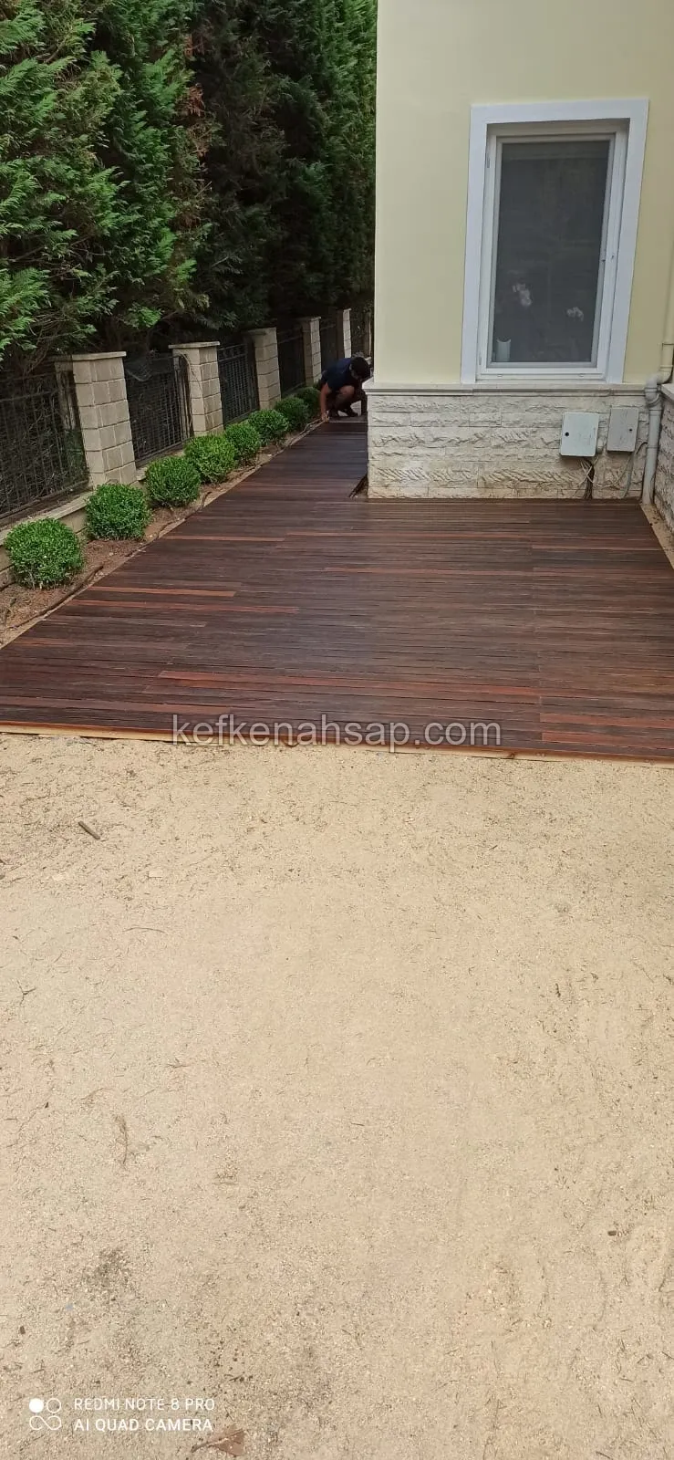 Sarı cepheli bir evin yanında, çit boyunca uzanan koyu kahverengi ahşap deck; bir usta zeminin ucunda çalışıyor.