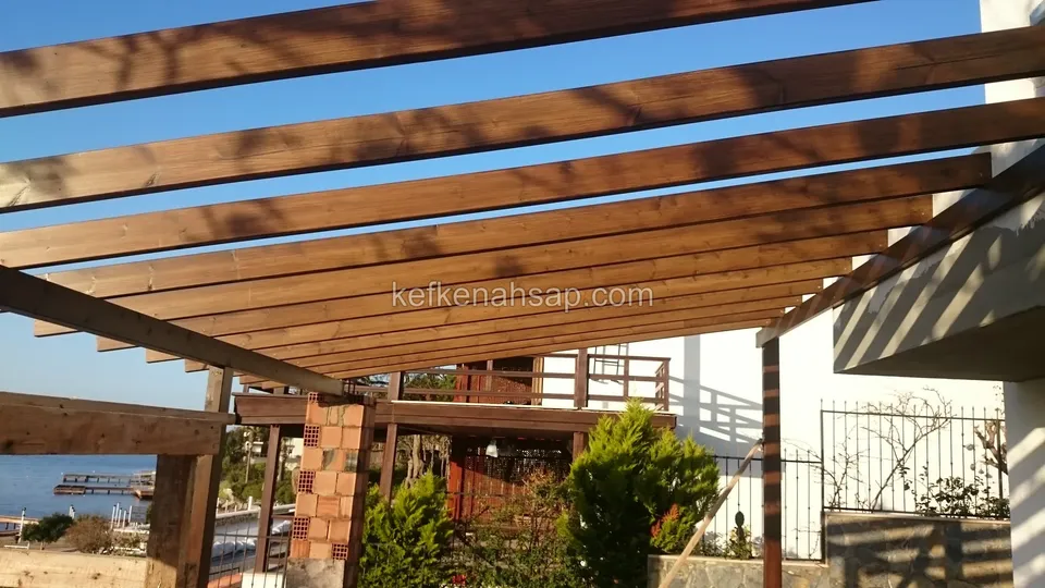 Villanın yanında vernikli pergola kirişleri, tuğla kolon, çalılık bahçe ve kenarda deniz