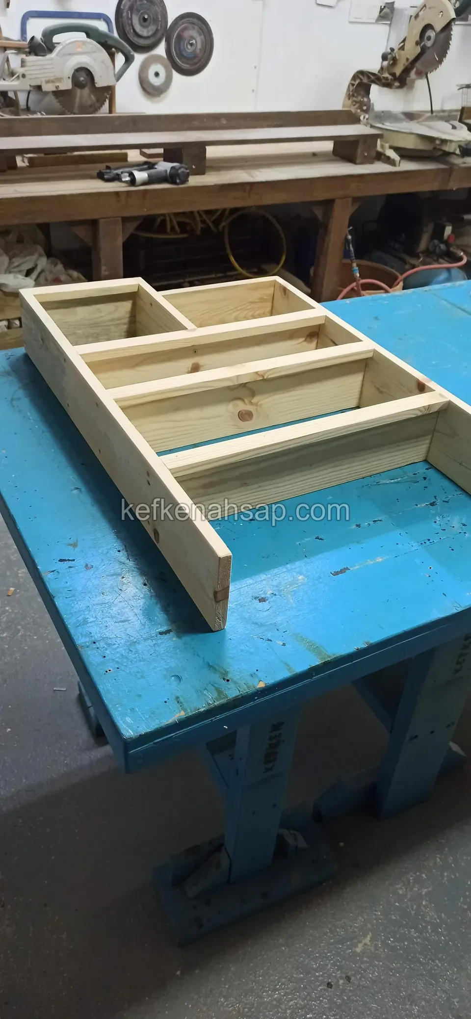 Ein Holzregalkorpus mit Fächern auf der Seite liegend auf einer blauen Werkbank, dahinter die Werkbank aus dicken Brettern