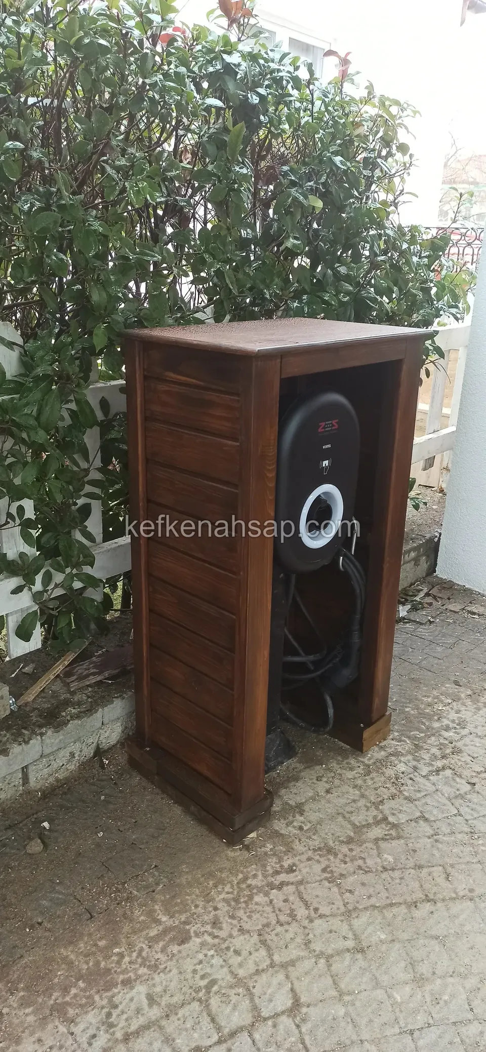 Çalıların önünde, parke taşlı zeminde duran, yan yüzü yatay çıtalı ve çatısı eğimsiz koyu ahşap kabin, içinde bir cihaz