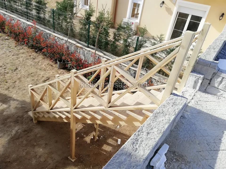 Seitenansicht einer Holztreppe mit Kreuzstreben-Geländer, dahinter Steinmauer, rote Sträucher und ein Haus