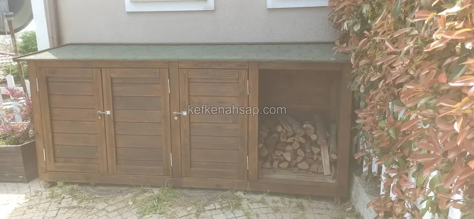 Beyaz duvar dibinde yatay tahtalı ahşap dolap, iki kapaklı kısım kapalı, sağ bölme açık ve içi odun dolu, çatı yeşil kaplı