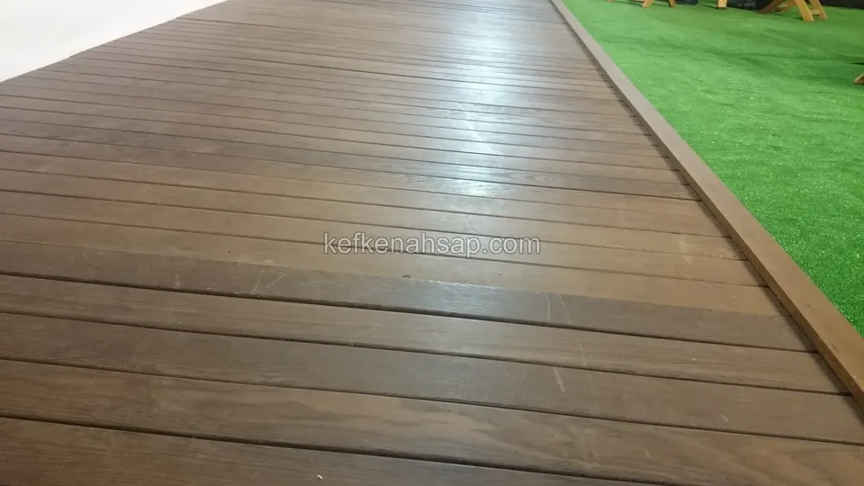 Yapay çimle sınırlanan, koyu kahve tonlu ve cilalı deck yüzeyi, kenarda sıralı bir bordür tahtası