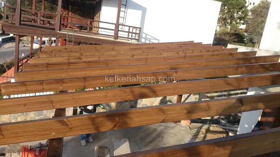 Pergola beams under construction with a house behind that has a timber-railed balcony, and a stone wall