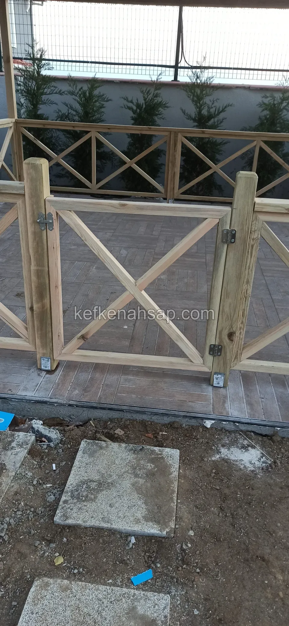 Tuğla döşemeli alanda kurulmakta olan X çaprazlı ahşap çit ve kapı bölümü; ön tarafta toprak ve beton plakalar