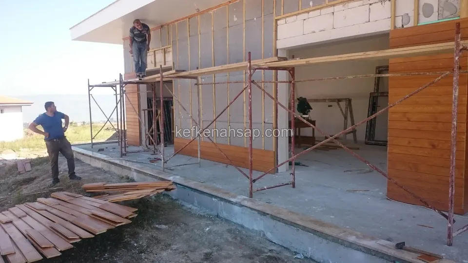 Yarısı tamamlanmış ahşap cephe kaplaması; iskelede çalışan biri, yerde kaplama tahtaları yığılı