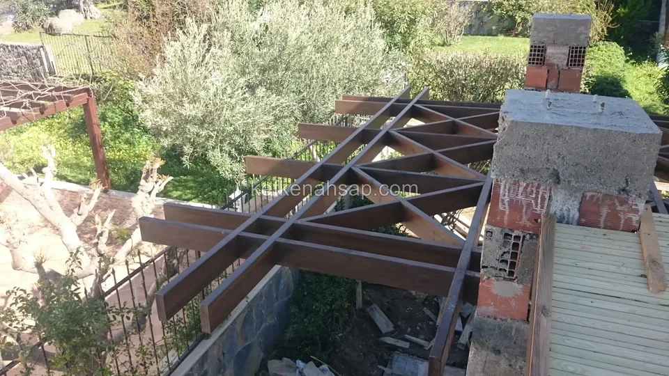Tamamlanmamış üçgen desenli pergola kafesi, yanında tuğla ve beton kolon, ahşap geçit ve zeytin ağaçları