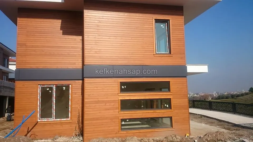 Yatay ahşap cephe kaplamalı iki katlı ev, ortada koyu gri bant, pencere kasaları ahşap, önünde inşaat toprağı