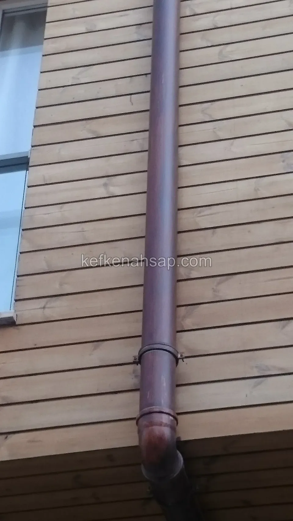 Açık kahve yatay ahşap cephe kaplamasında dikey inen yağmur borusu ve altta çıkma bir bölüm, solda cam yüzey