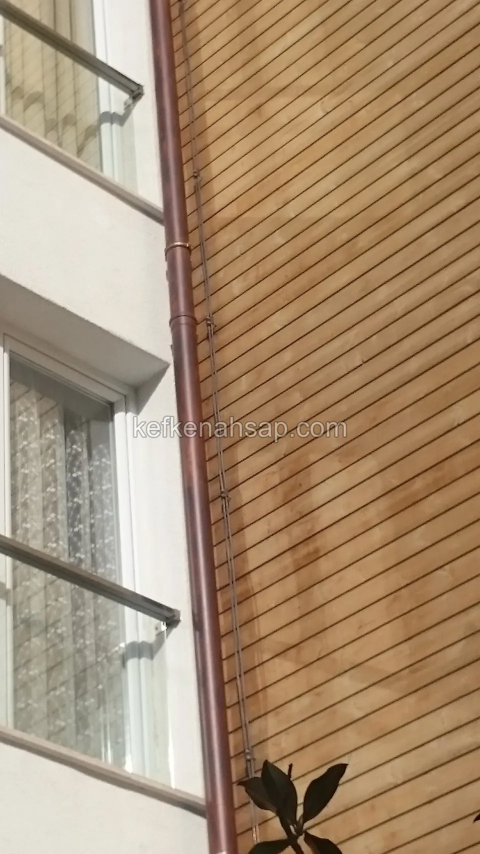 Açık tonlu yatay ahşap kaplamalı cephe, solda cam tuğlalı pencereler ve beyaz duvar, kenarda bakır renkli yağmur borusu
