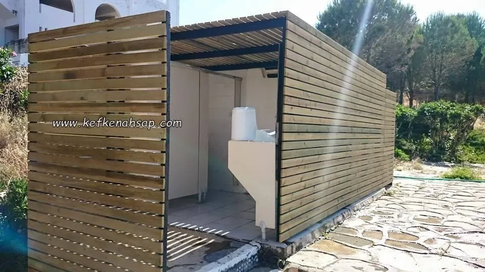 Yatay ahşap çıtalarla kaplanmış, üstü çıtalı örtülü açık girişli WC ve lavabo kabini; önünde taş döşeli zemin var