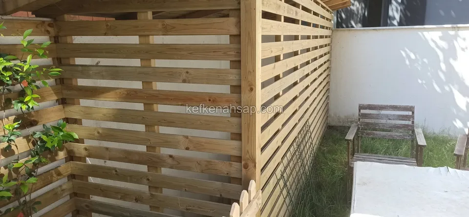 Beyaz duvarlı bahçe köşesinde yatay çıtalı ahşap paravan ve önünde eski bir ahşap sandalye