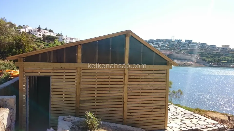 Yatay çıtalı kaplamalı, koyu üçgen üst bölümlü kabin; yanında taş döşeli kıyı ve arkada deniz